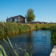 Lille hus kaldet Berkel ved en fredelig dam omgivet af rør og træer under en klar blå himmel.