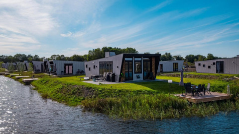 Moderne Just Nature Lodges am Seeufer mit Grünfläche, Terrasse und Wasser bei sonnigem Wetter.