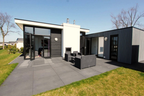 Lodge moderne L-Cube avec façade noire et grise, grandes fenêtres, terrasse et mobilier extérieur.