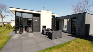 Moderne L-Cube lodge met zwarte en grijze gevel, grote ramen, openslaande deuren en terrasmeubilair.