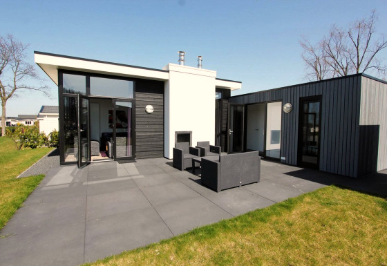Lodge moderne L-Cube avec façade noire et grise, grandes fenêtres, terrasse et mobilier extérieur.