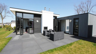 Strakke L-Cube lodge met donkere gevel, grote raampartijen en gezellig terras met tuinmeubilair.