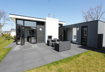 Strakke L-Cube lodge met donkere gevel, grote raampartijen en gezellig terras met tuinmeubilair.
