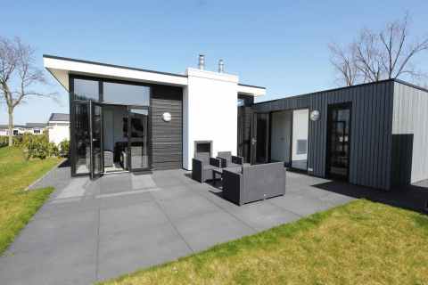 Strakke L-Cube lodge met donkere gevel, grote raampartijen en gezellig terras met tuinmeubilair.