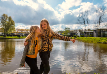 Dos niñas felices posan junto al lago frente al alojamiento L-Cube en Spaarnwoude, Países Bajos, día soleado.