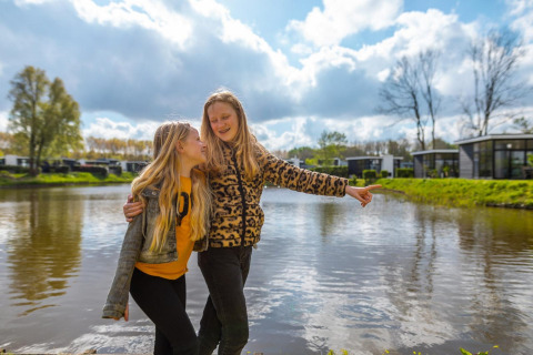 Dos niñas felices posan junto al lago frente al alojamiento L-Cube en Spaarnwoude, Países Bajos, día soleado.