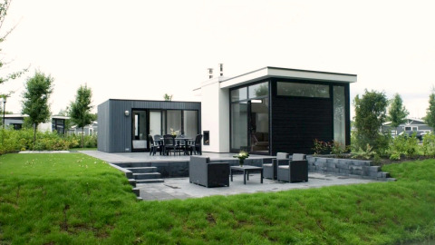 Moderne L-Cube-lodge met groot terras, tuinmeubels en groene tuin in een rustige, natuurlijke omgeving.