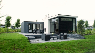 Moderne L-Cube-lodge met groot terras, tuinmeubels en groene tuin in een rustige, natuurlijke omgeving.