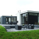 Moderne L-Cube lodge med stor terrasse, havemøbler og grøn have i fredelige omgivelser.