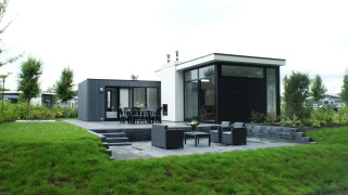 Moderne L-Cube lodge med stor terrasse, havemøbler og grøn have i fredelige omgivelser.