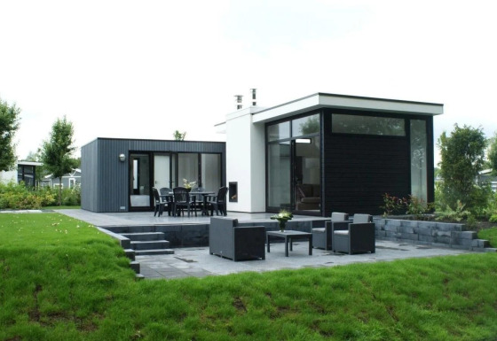 Moderne L-Cube lodge med stor terrasse, havemøbler og grøn have i fredelige omgivelser.