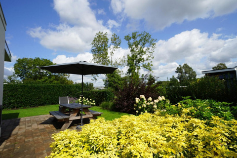 Jardín con flores, arbustos verdes y terraza con mesa de picnic y sombrilla en Cube Maximaal en Spaarnwoude.