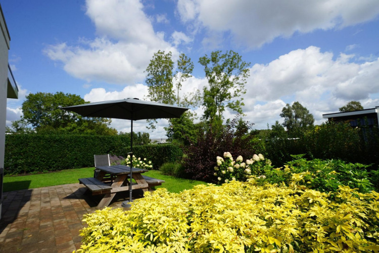 Jardín con flores, arbustos verdes y terraza con mesa de picnic y sombrilla en Cube Maximaal en Spaarnwoude.