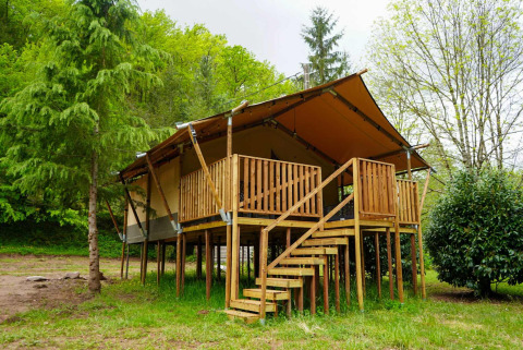 Tenda safari con servizi igienici su palafitte, circondata da alberi, con veranda e scala in legno.