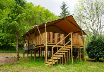 Tenda safari con servizi igienici su palafitte, circondata da alberi, con veranda e scala in legno.