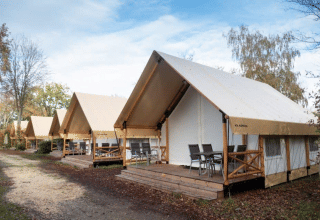 Rij moderne safaritenten met houten veranda's en tuinmeubilair op een rustige, groene campinglocatie.