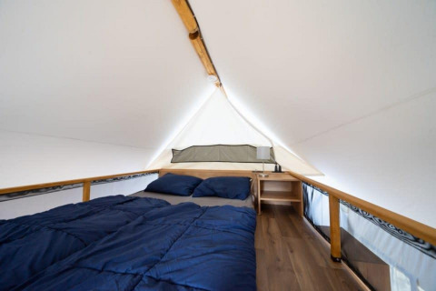 Vista interior de una tienda safari con cama doble, ropa de cama azul, suelo de madera y mesita con lámpara.