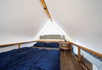 Interior view of a safari tent with a double bed, blue bedding, wooden floor, and a nightstand lamp.