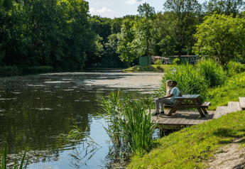 Dos personas sentadas en una mesa junto al lago en un glamping, rodeados de vegetación y árboles.