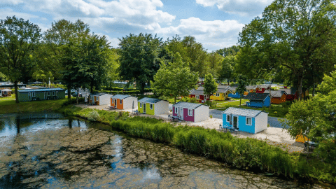 Cabañas de glamping coloridas junto a un lago tranquilo, rodeadas de árboles y cielo azul con nubes.