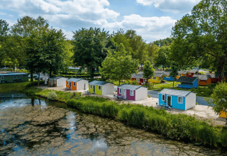 Cabañas de glamping coloridas junto a un lago tranquilo, rodeadas de árboles y cielo azul con nubes.