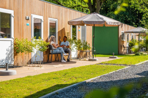Escena exterior en una cabaña en Het Amsterdamse Bos, Países Bajos, con dos personas sentadas afuera al sol.