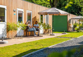 Escena exterior en una cabaña en Het Amsterdamse Bos, Países Bajos, con dos personas sentadas afuera al sol.