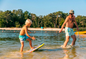 Ein Mann zieht ein Kind auf einem Skimboard im flachen Wasser am Strand beim Glamping-Urlaub.