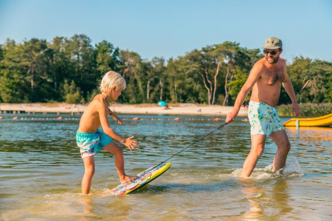 Ein Mann zieht ein Kind auf einem Skimboard im flachen Wasser am Strand beim Glamping-Urlaub.