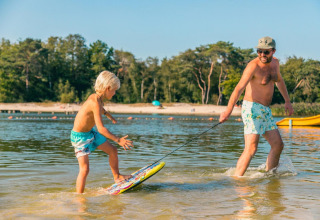 Een man trekt een kind op een skimboard in ondiep water aan het strand tijdens een glampingverblijf.