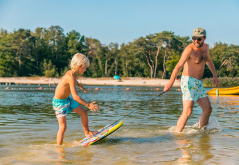 Ein Mann zieht ein Kind auf einem Skimboard im flachen Wasser am Strand beim Glamping-Urlaub.