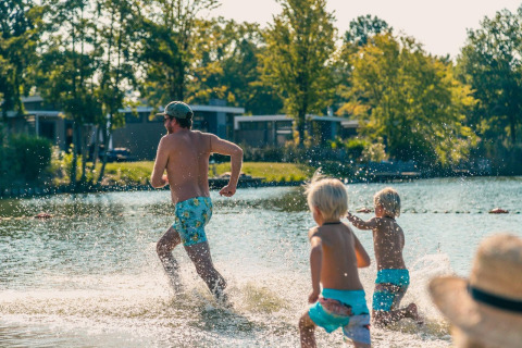 Famille courant et jouant dans l'eau peu profonde d’un glamping, avec cabanes et arbres en arrière-plan.
