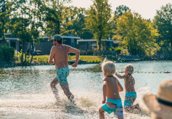 En familie løber og leger i vandet ved et glamping-sted, omgivet af træer og små hytter i baggrunden.