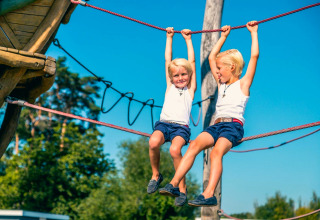 Deux enfants jouent sur une corde d’escalade en plein air dans un hébergement glamping sous ciel bleu.