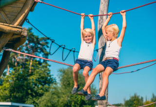 Zwei Kinder spielen an einem Kletterseil im Freien auf einem Glamping-Platz bei strahlendem Wetter.