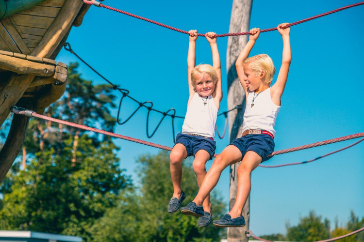 Zwei Kinder spielen an einem Kletterseil im Freien auf einem Glamping-Platz bei strahlendem Wetter.