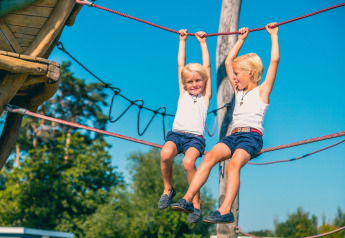 Dos niños juegan en una cuerda para escalar al aire libre bajo el cielo azul en un alojamiento glamping.