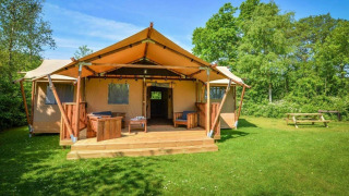 Glamping accommodatie met canvas tent, houten terras en zithoek, omgeven door gras en bos.