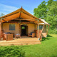Glampingindkvartering med teltstruktur, træterrasse og siddepladser omgivet af frodige grønne træer.