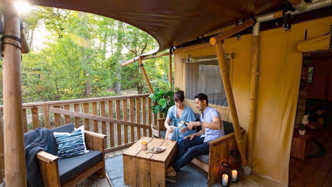 Un couple savoure des boissons sur une terrasse en bois confortable entourée d’arbres dans un glamping chic.