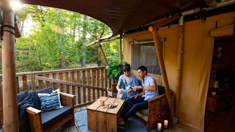Een koppel geniet van een drankje op een gezellig houten terras bij een glampingaccommodatie in het bos.