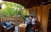 Un couple savoure des boissons sur une terrasse en bois confortable entourée d’arbres dans un glamping chic.