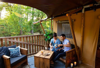 Una coppia si gode una bevanda su una terrazza di legno immersa nel verde in un elegante glamping.