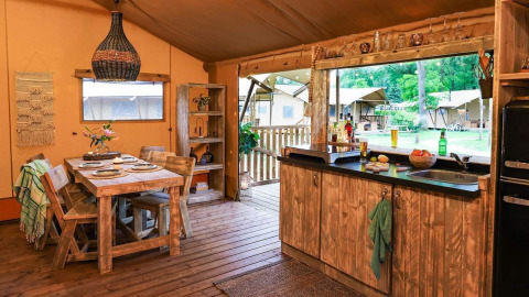 Gezellige glampingaccommodatie met houten meubels, eettafel en open keuken met zicht op de natuur.