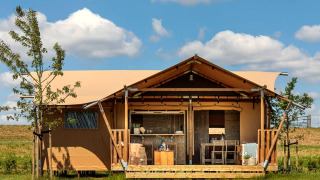 Tienda glamping con terraza de madera y muebles exteriores, rodeada de naturaleza y cielo azul.