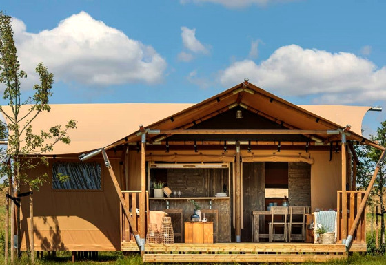 Tente de glamping avec véranda en bois et salon de jardin, entourée de nature sous un ciel bleu.