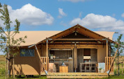 Tente de glamping avec véranda en bois et salon de jardin, entourée de nature sous un ciel bleu.