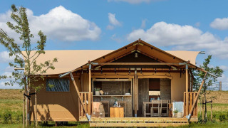 Glampingtent met houten terras en tuinmeubelen, gelegen in de natuur onder een heldere blauwe lucht.
