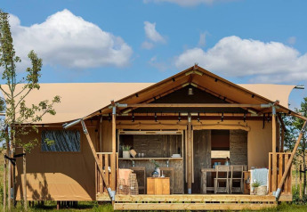 Tente de glamping avec véranda en bois et salon de jardin, entourée de nature sous un ciel bleu.