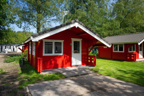 Cabaña roja con puerta y ventanas blancas, situada en una zona verde y arbolada bajo la luz del sol.